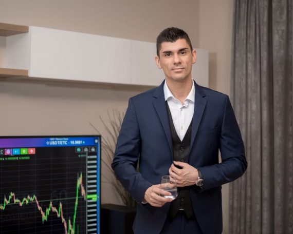 A businessman in a suit stands indoors with a stock market graph on a screen.